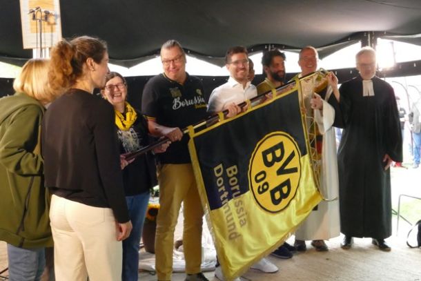 Mehrere Menschen stehen zusammen und halten eine große schwarz-gelbe Fahne mit dem Logo und Schriftzug von Borussia Dortmund (BVB 09). Einige tragen Fanartikel in den Vereinsfarben. Rechts im Bild stehen zwei Geistliche, einer im weißen Gewand mit Stola, der andere im schwarzen Talar. Alle befinden sich unter einem Zelt
